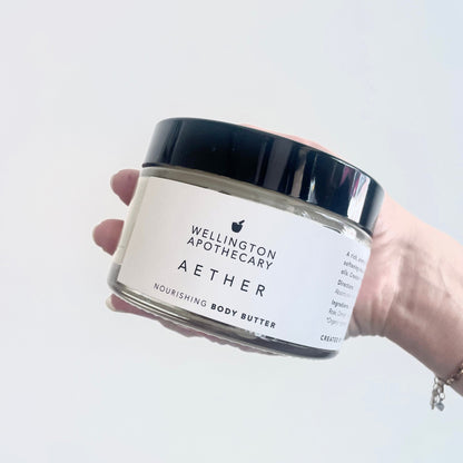 Glass jar of aether nourishing body butter held in hand in front of white background.