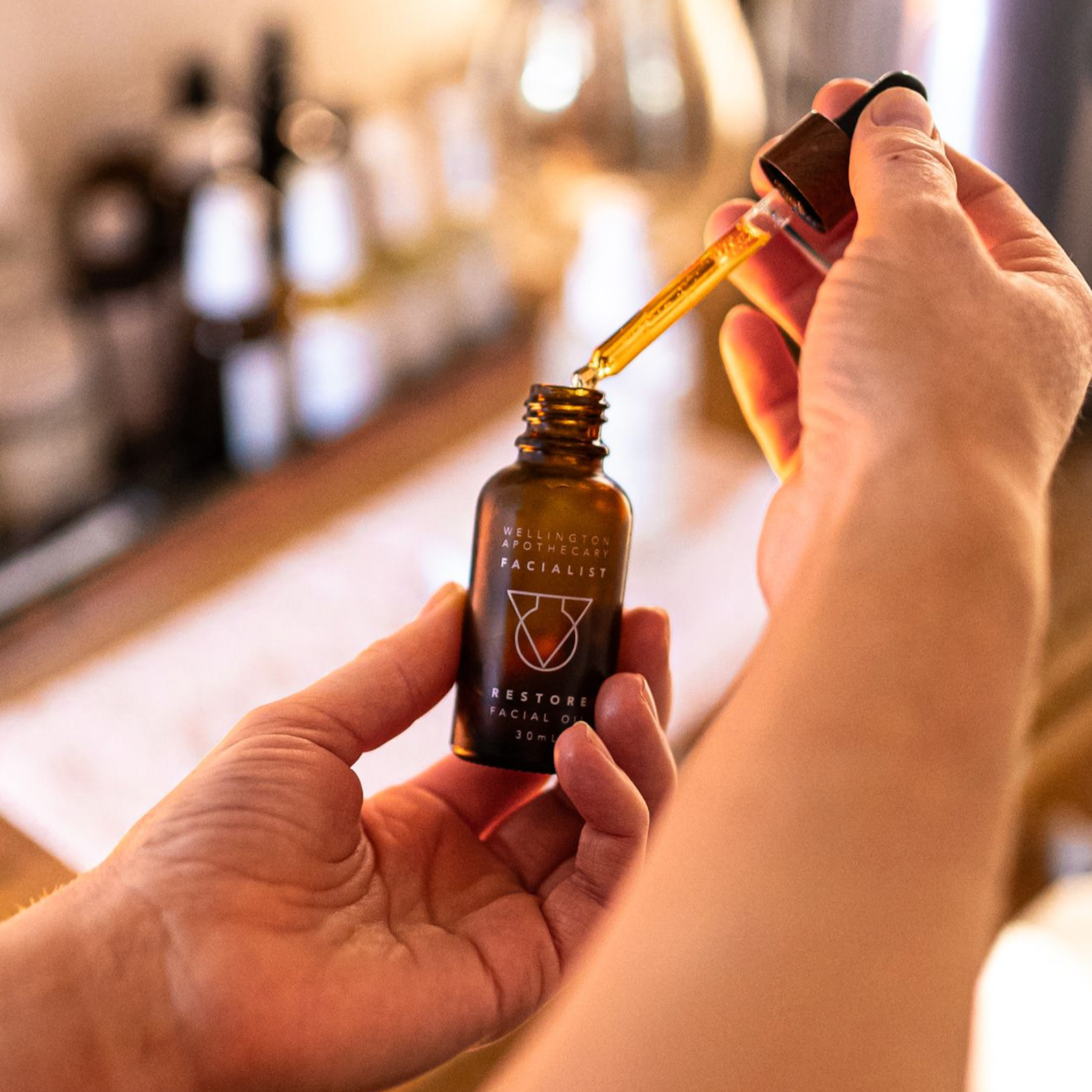 Hand holding amber glass bottle of restore facial oil, other hand holding glass dropper full of golden coloured oil, with blurred therapy room background.