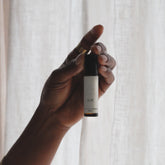 An elegant hand holding the Air roll on perfume blend in soft, dim lighting in front of a sheer natural coloured curtain. 