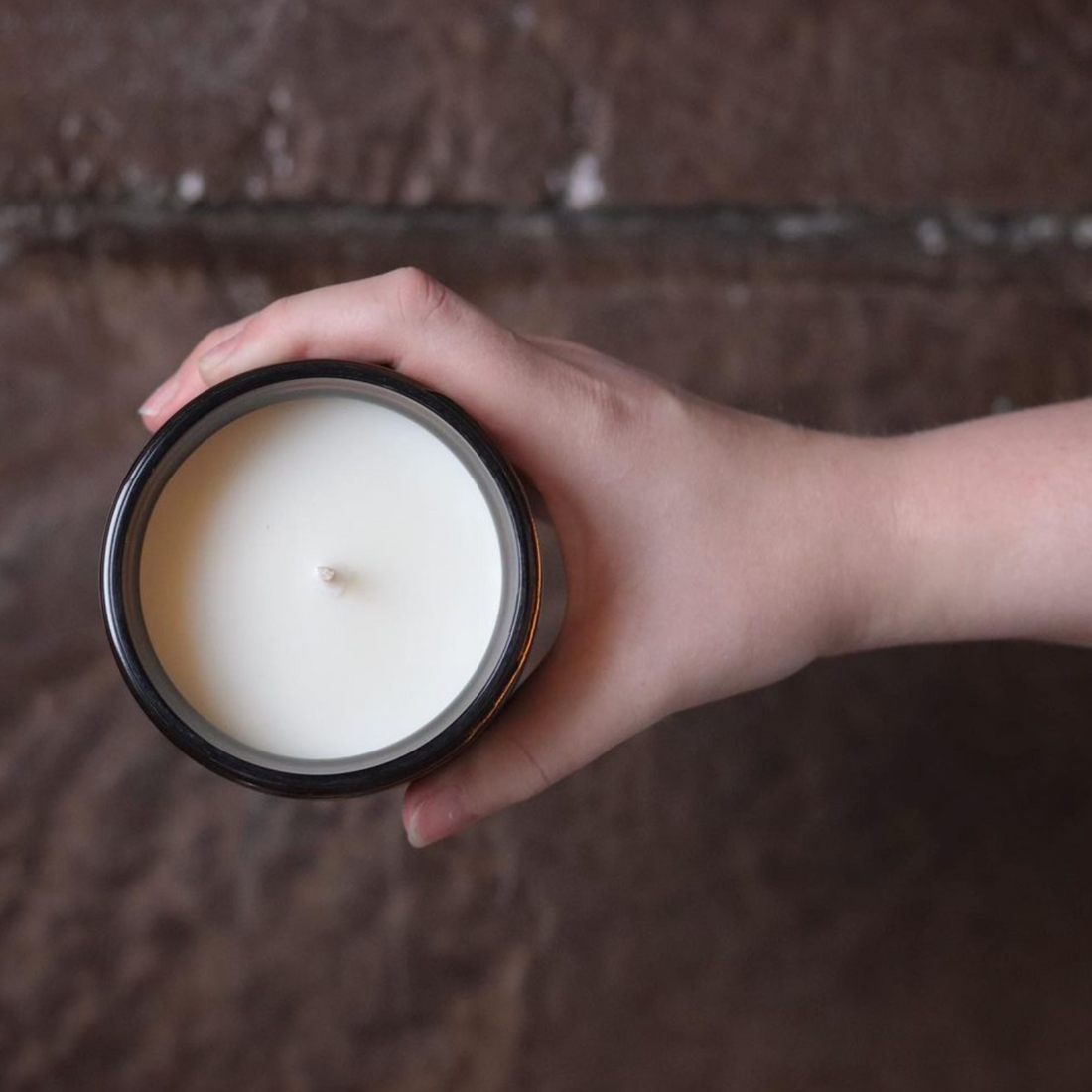 An aerial view of an open soy wax candle in an amber glass jar, held by a hand in front of a brown brick surface.