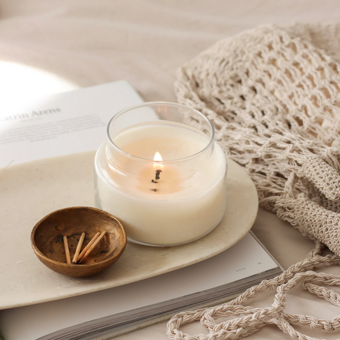 Glass apothecary jar with a lit coconut wax candle on a beige earthenware dish, a brass bowl of matches, an open book and a natural fibre woven bag. 