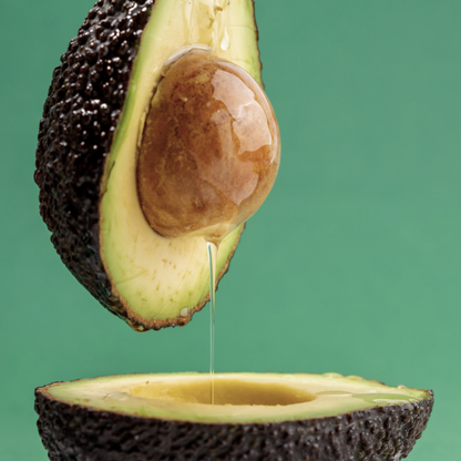 An avocado cut in half, oil dripping from the half held above the other, with a green background. 