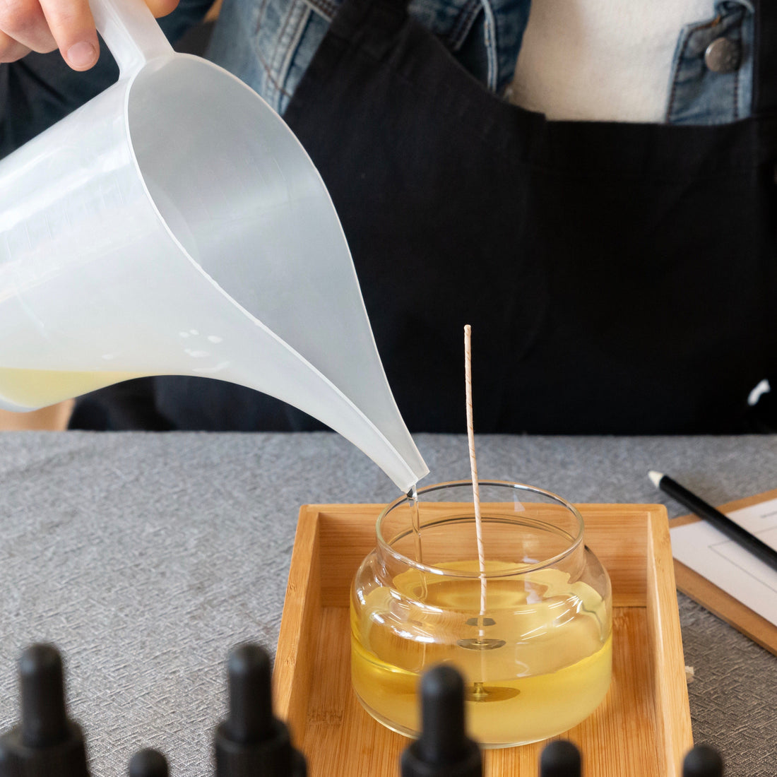 Person wearing black apron pouring melted wax from a jug into a glass apothecary jar.