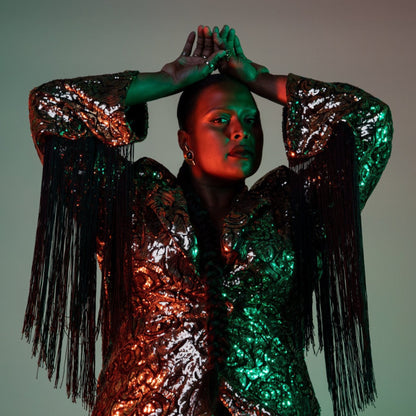 Portrait of Deva in dark light wearing a shimmering pink and green jacket with long black tassels. 