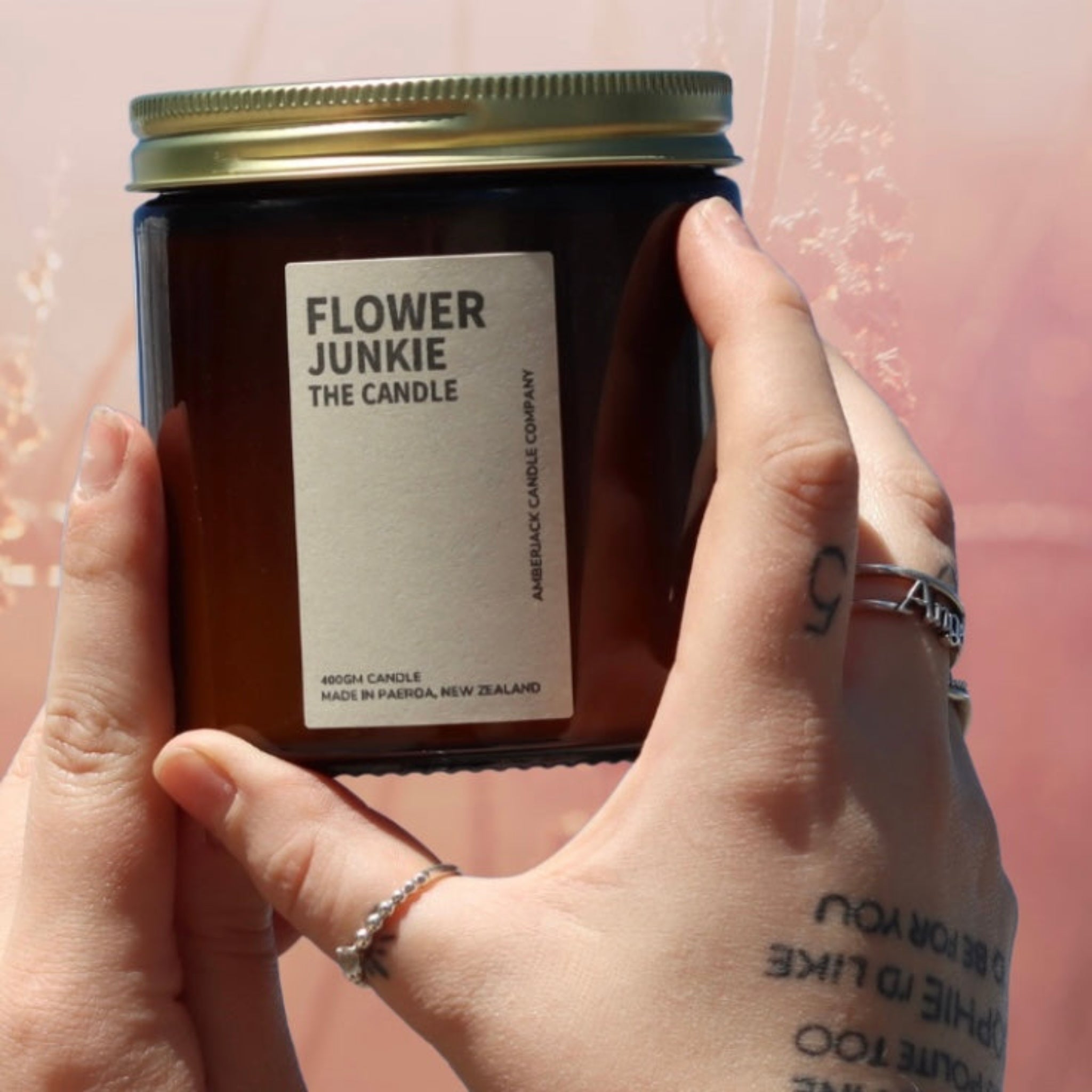 Flower junkie candle in amber glass jar with brass lid held in hands with tattoos and rings in front of soft pink floral background. 