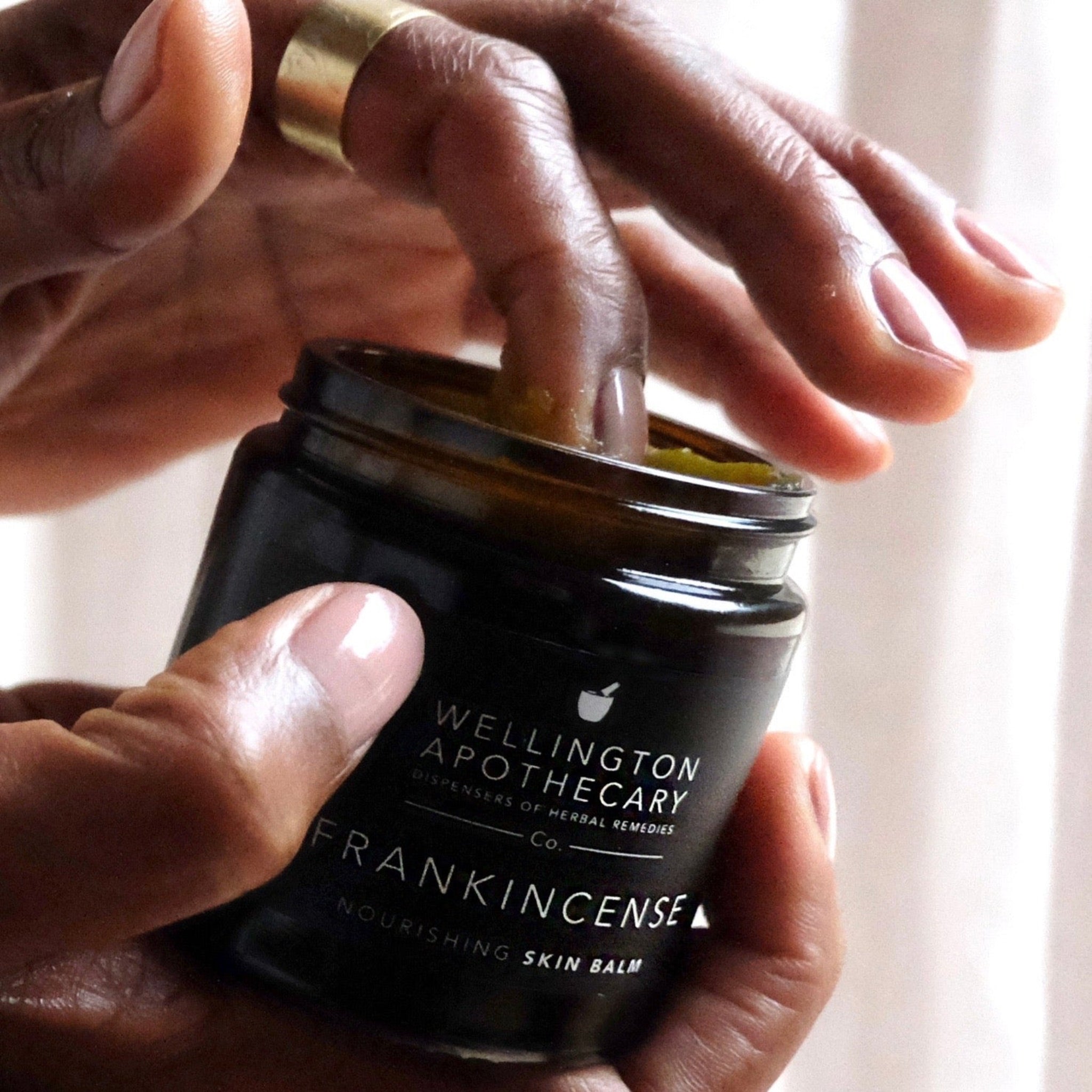 Open glass jar of frankincense balm, held in one hand while the other digs a finger in.