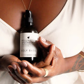 Woman holding a bottle of Holy Basil cleansing gel in front of her chest, wearing white silk and gold jewellery. 