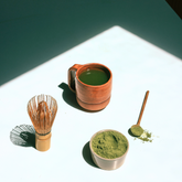 A cup of matcha tea, a matcha whisk, a spoonful of matcha powder and a small bowl full of matcha powder on a white bench. 