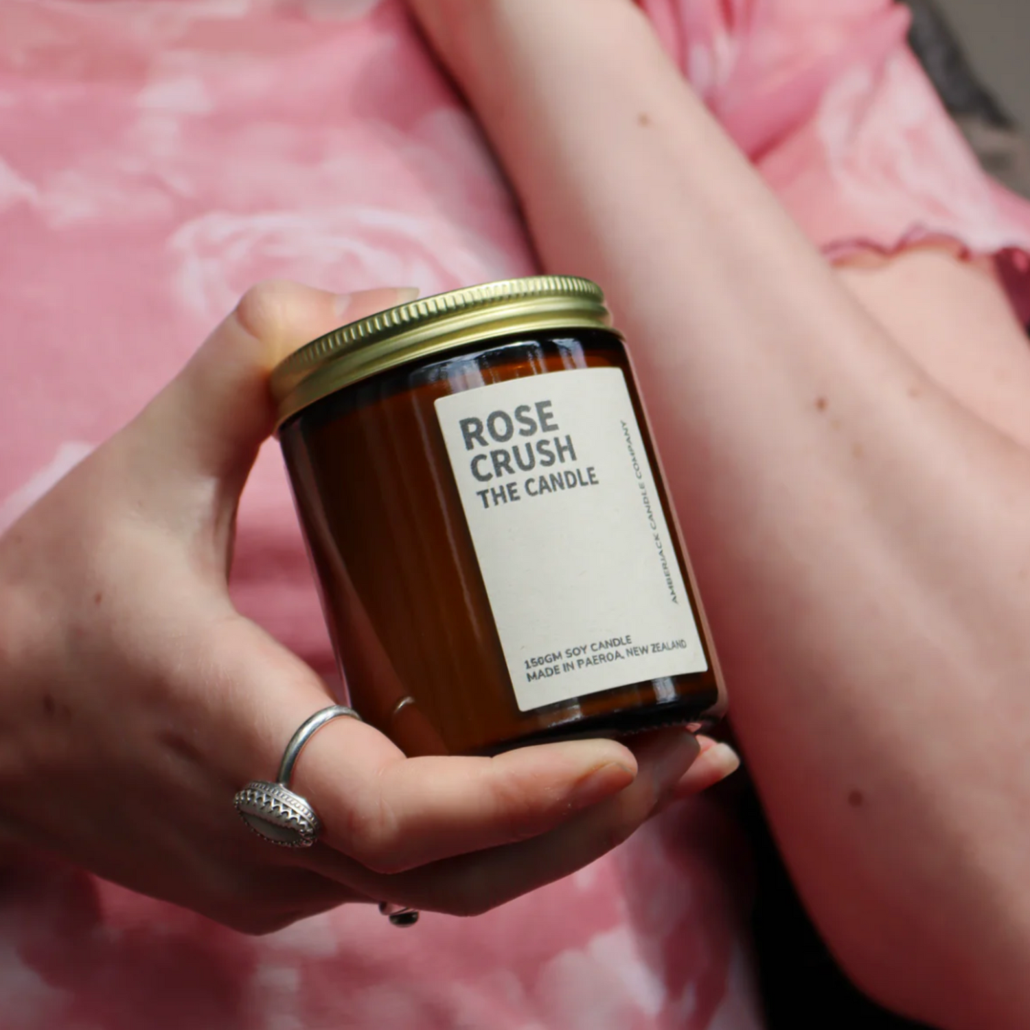 Amber glass jar with brass lid containing rose crush candle, held by person in a rose printed top. 