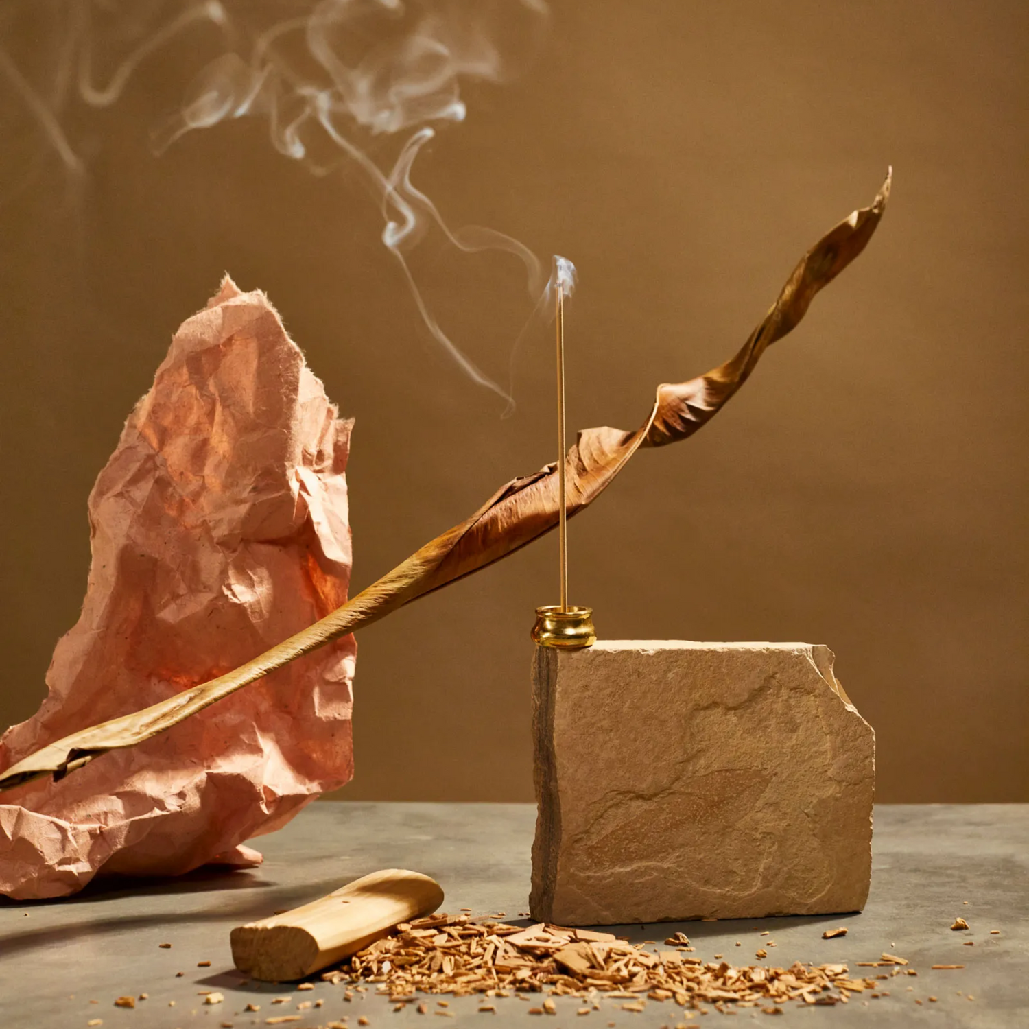 Sandalwood incense burning in brass holder a top of a block of stone, with woods chips, a dried leaf and crumpled brown paper.