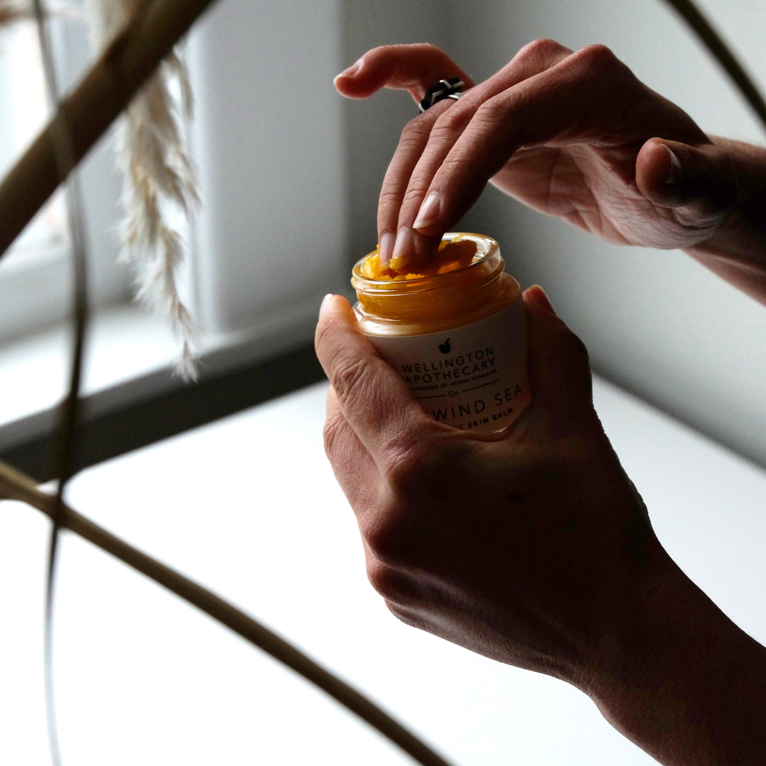 Hand holding jar of sun wind sea balm while pressing fingers in, softly lit space with wisps of seagrass framing the image. 