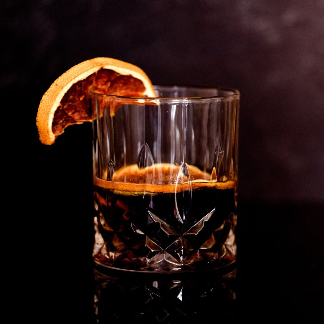 A crystal glass of dark whiskey with a slice of dark dehydrated orange with a dark aubergine background.