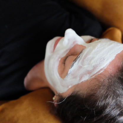 Woman lying on mustard coloured couch with eyes closed and ivory clay mask on her face.