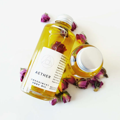 Clear glass bottles of golden Aether body oil infused with roses and surrounded by dried pink roses. 