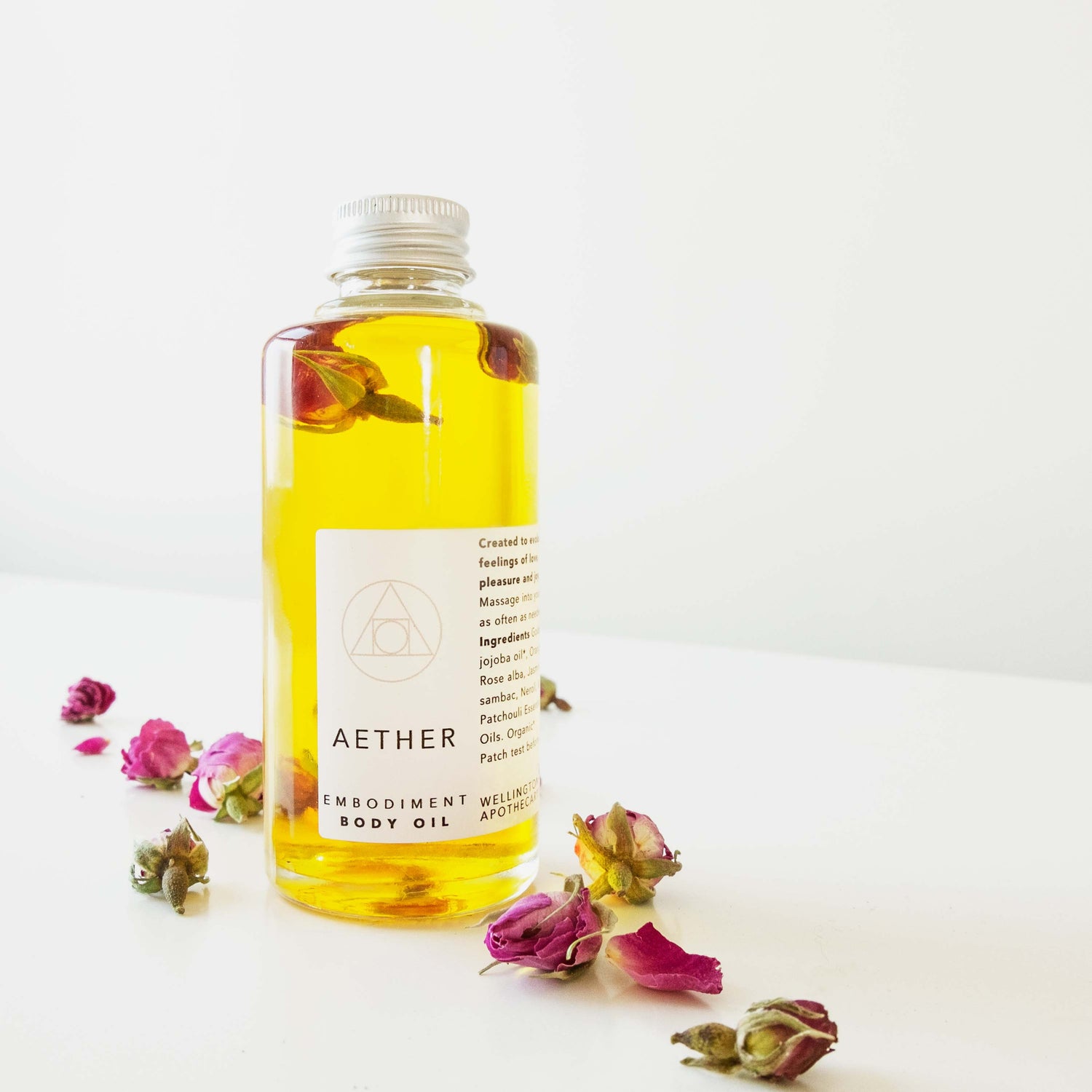 Clear glass bottle of golden Aether body oil infused with roses and surrounded by dried pink roses. 