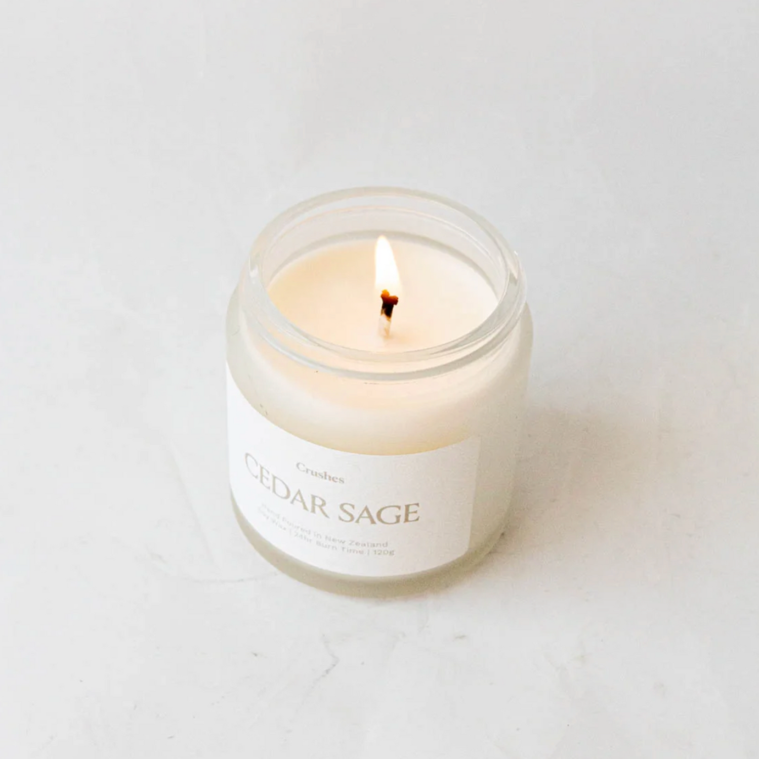 Clear glass jar containing cedar sage candle alight on white background. 