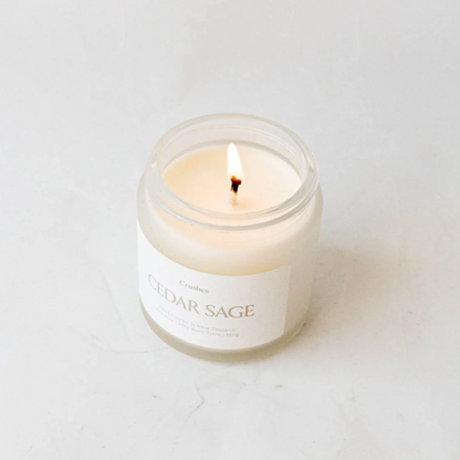 Clear glass jar containing cedar sage candle alight on white background. 