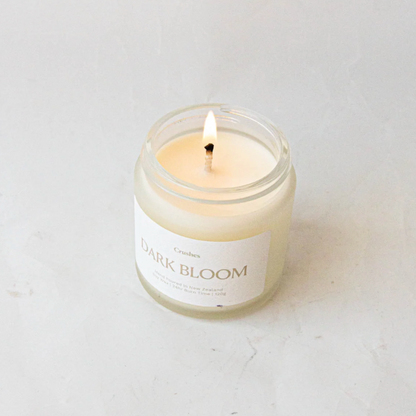 Clear glass jar containing dark bloom candle alight  on white background. 