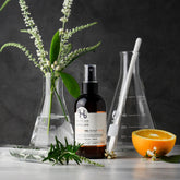 Spray bottle of Holistic Hair scalp spray on bench with orange, rosemary, neroli and conical flasks. 