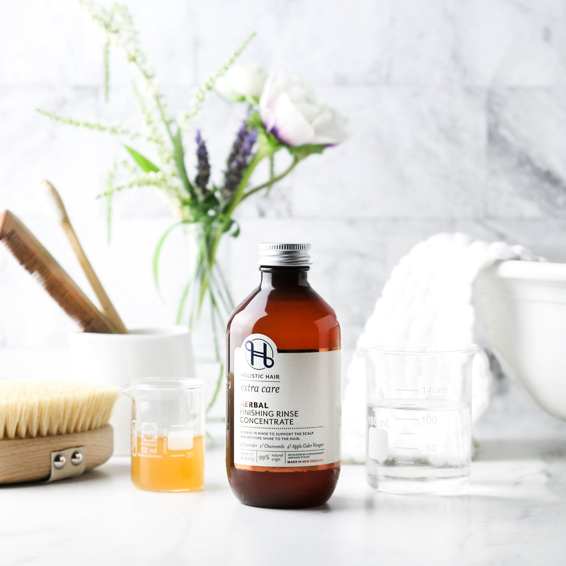 Bottle of Herbal Hair Rinse concentrate on bench with body brush, wooden comb, a towel, some flowers and a beaker of apple cider vinegar. 