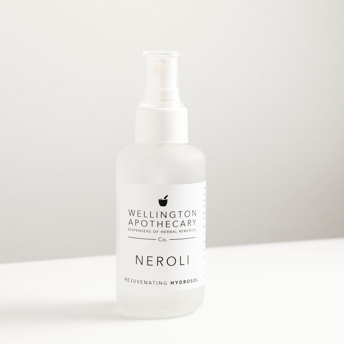 Frosted white glass bottle filled with neroli hydrosol, on white bench and soft grey background.