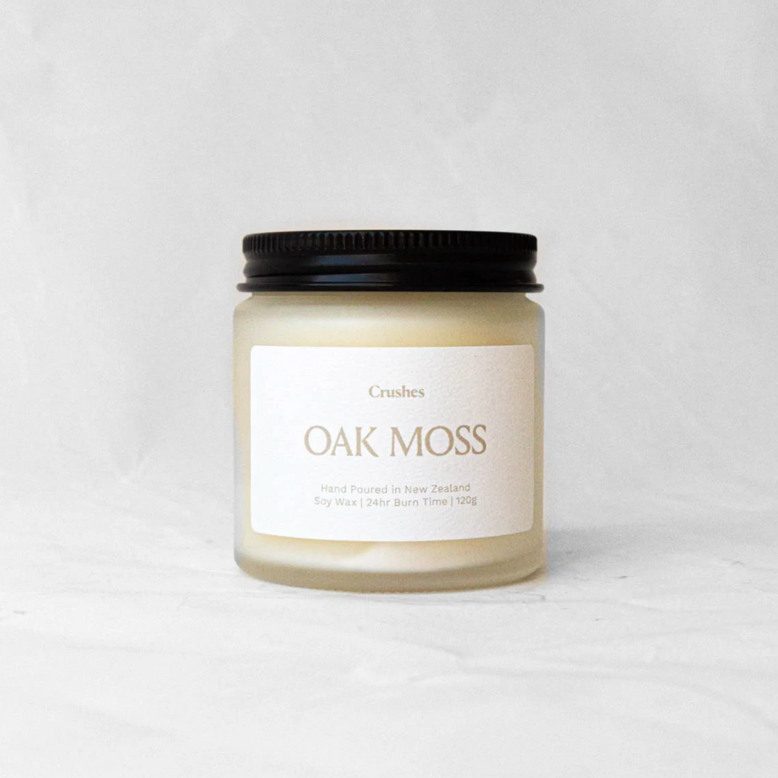 Clear glass jar with black metal lid containing oak moss candle on white background. 