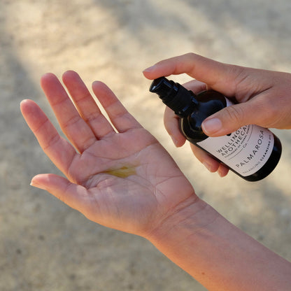 Palmarosa cleansing oil being dispensed onto open palm with sandy sunlit background. 