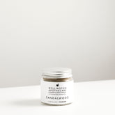 Glass jar with aluminium lid on white bench filled with sandalwood exfoliant powder. 