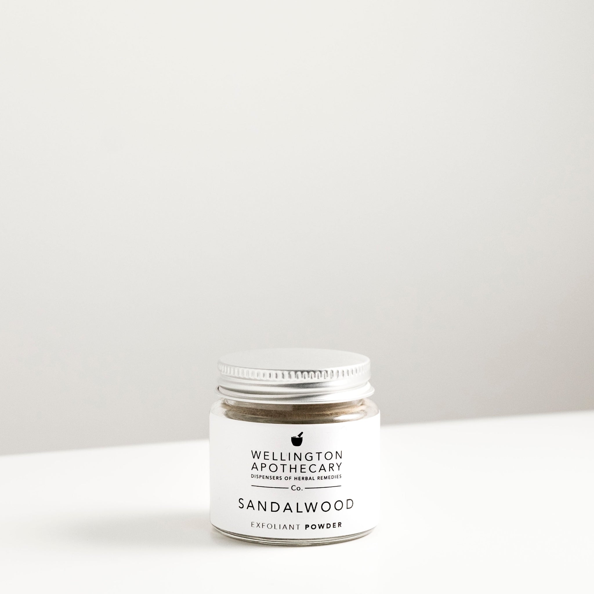 Glass jar with aluminium lid on white bench filled with sandalwood exfoliant powder. 