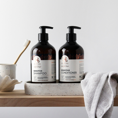 500mL bottles of Holistic Hair Sensitive shampoo and conditioner on ceramic dish in bathroom setting with a hand towel, shell and ceramic cup holding a toothbrush. 