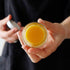 Open jar of golden yellow balm held in hand with black clothing background. 