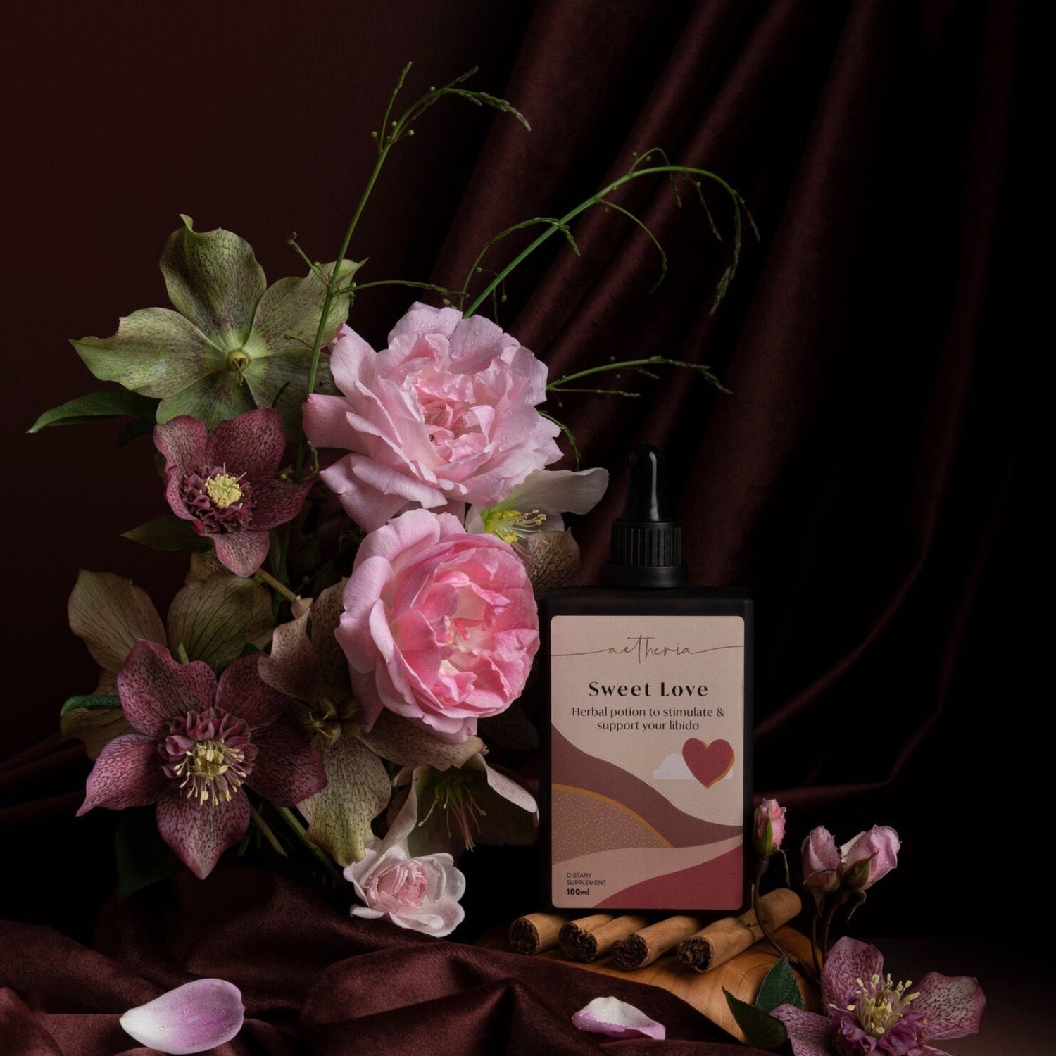 A bottle of sweet love tincture in a velvety maroon setting with roses, hellebore flowers and cinnamon quills.